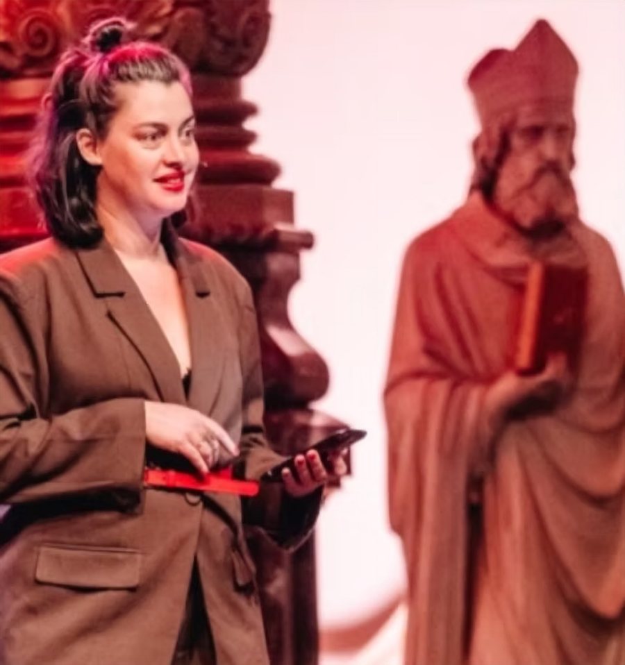 Kim Triesscheijn op het podium, met rode lippen en een bruin colbert, tijdens een spoken word optreden